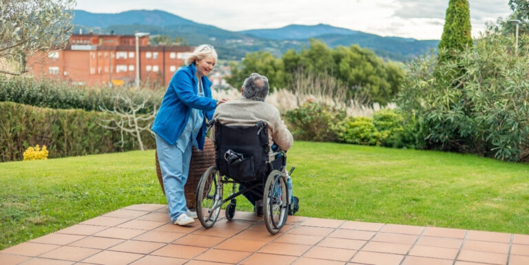Female Nurse with an Elderly Resident in a Wheelchair Outdoors at a Memory Care Facility